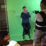Ryan sitting on a stool in front of green screen while Wes films with camera.